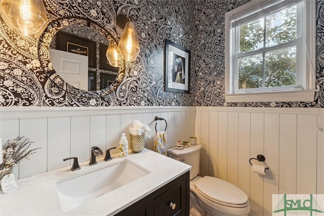 half bathroom with wainscoting, vanity, toilet, and wallpapered walls