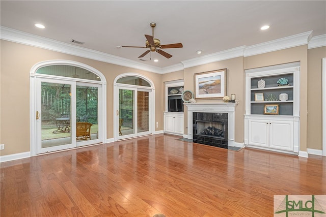 unfurnished living room featuring ceiling fan, built in features, crown molding, a fireplace, and light wood-type flooring