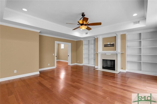 unfurnished living room with ceiling fan, built in shelves, hardwood / wood-style flooring, a tray ceiling, and crown molding