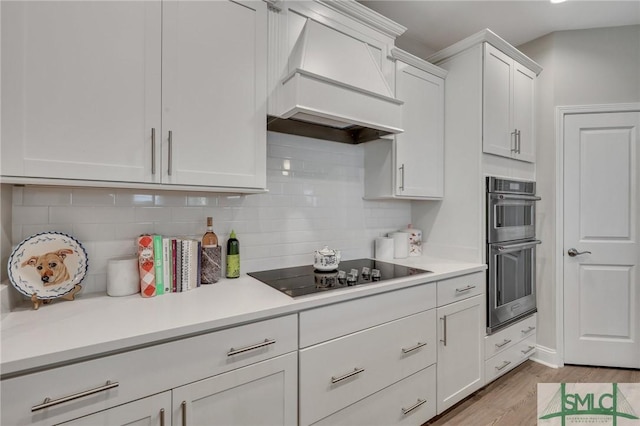 kitchen featuring custom exhaust hood, double oven, black electric stovetop, light hardwood / wood-style floors, and white cabinets