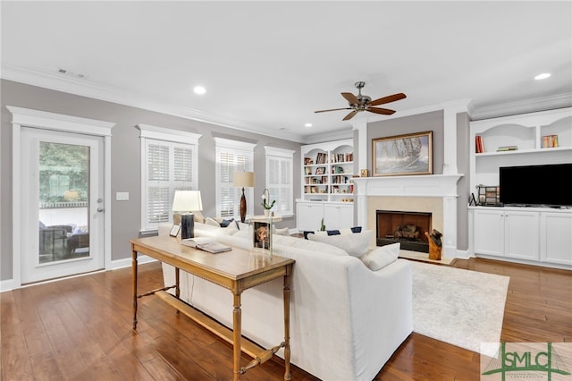 living room featuring built in shelves, ornamental molding, ceiling fan, a premium fireplace, and hardwood / wood-style floors