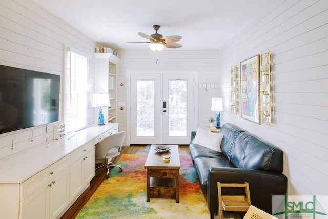 living room with built in desk, wood walls, ceiling fan, french doors, and hardwood / wood-style flooring