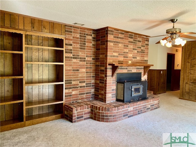 unfurnished living room with ceiling fan, wood walls, carpet flooring, a wood stove, and a textured ceiling