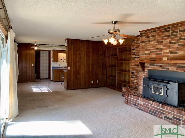 unfurnished living room with wood walls, a wood stove, a textured ceiling, ceiling fan, and light colored carpet