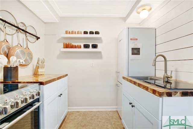 kitchen with stainless steel range with electric cooktop, wooden counters, sink, light tile patterned flooring, and white cabinetry