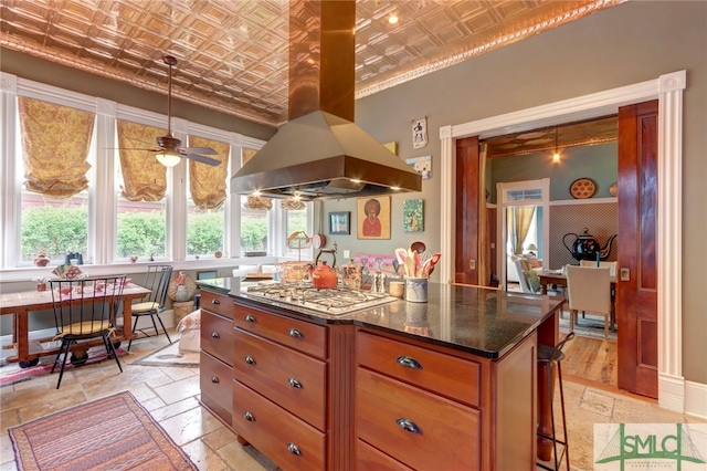 kitchen featuring island exhaust hood, ceiling fan, decorative light fixtures, stainless steel gas stovetop, and dark stone counters