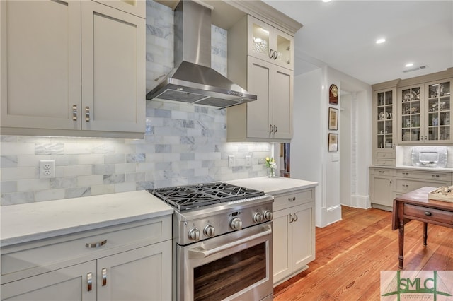 kitchen with wall chimney range hood, high end range, backsplash, and light wood-type flooring