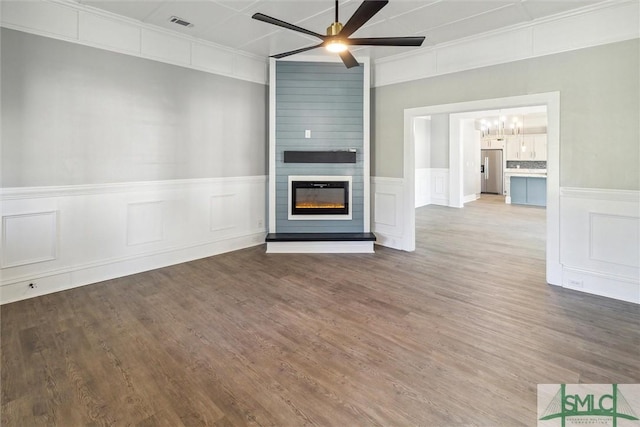 unfurnished living room with a large fireplace, a wainscoted wall, a decorative wall, ceiling fan with notable chandelier, and wood finished floors