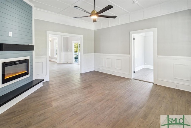 unfurnished living room with a decorative wall, ceiling fan, a glass covered fireplace, a wainscoted wall, and wood finished floors