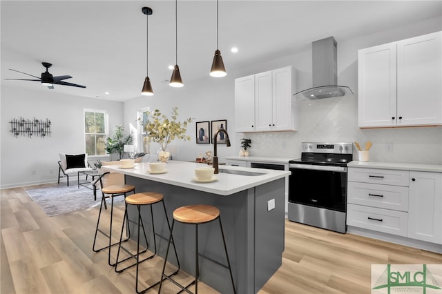 kitchen with white cabinetry, stainless steel electric range oven, wall chimney exhaust hood, pendant lighting, and a kitchen island with sink