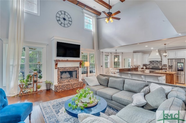 living room with beam ceiling, dark wood-type flooring, a fireplace, a towering ceiling, and ceiling fan