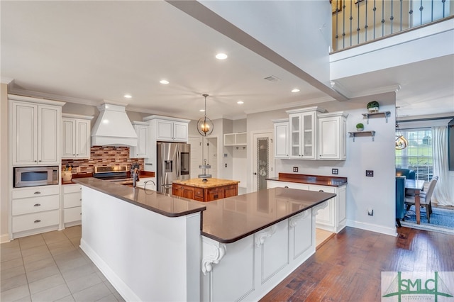 kitchen featuring custom range hood, appliances with stainless steel finishes, white cabinetry, and a kitchen island with sink