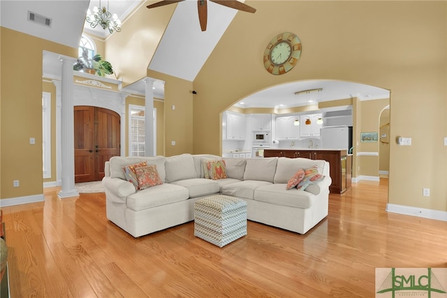 living room with ceiling fan with notable chandelier, ornate columns, light wood-type flooring, and high vaulted ceiling