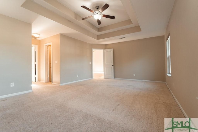 empty room with a raised ceiling, light carpet, ceiling fan, and baseboards