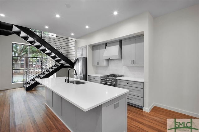 kitchen featuring gray cabinetry, appliances with stainless steel finishes, wood-type flooring, custom exhaust hood, and a kitchen island with sink