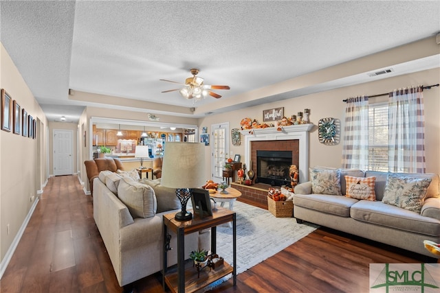 living room with ceiling fan, a textured ceiling, a tray ceiling, dark wood-type flooring, and a fireplace