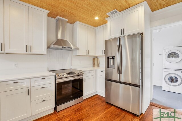 kitchen with hardwood / wood-style floors, white cabinetry, wall chimney exhaust hood, stainless steel appliances, and stacked washer and clothes dryer