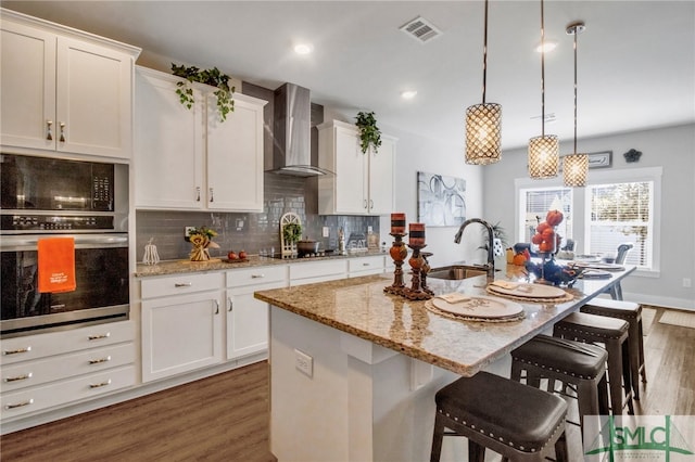 kitchen with sink, stainless steel oven, hanging light fixtures, wall chimney exhaust hood, and a center island with sink