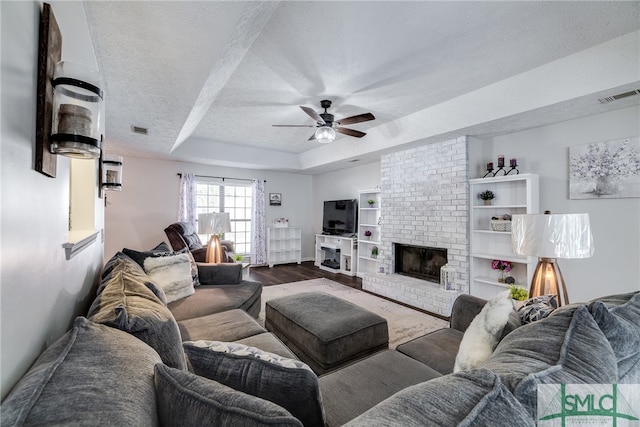 living room featuring a raised ceiling, a textured ceiling, a brick fireplace, ceiling fan, and hardwood / wood-style flooring