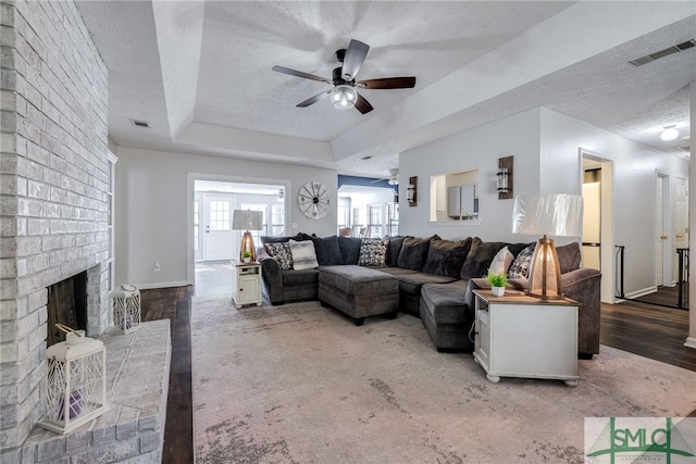 living room with a tray ceiling, hardwood / wood-style floors, a brick fireplace, a textured ceiling, and ceiling fan