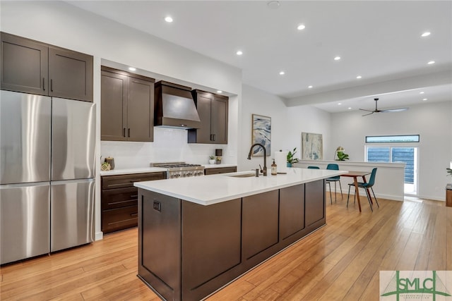 kitchen featuring stainless steel appliances, a center island with sink, sink, wall chimney range hood, and light hardwood / wood-style flooring