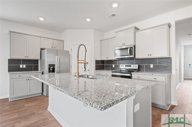 kitchen with sink, appliances with stainless steel finishes, backsplash, a kitchen island with sink, and light hardwood / wood-style flooring