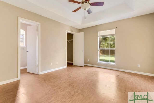 unfurnished bedroom with a tray ceiling, a spacious closet, ceiling fan, wood finished floors, and baseboards