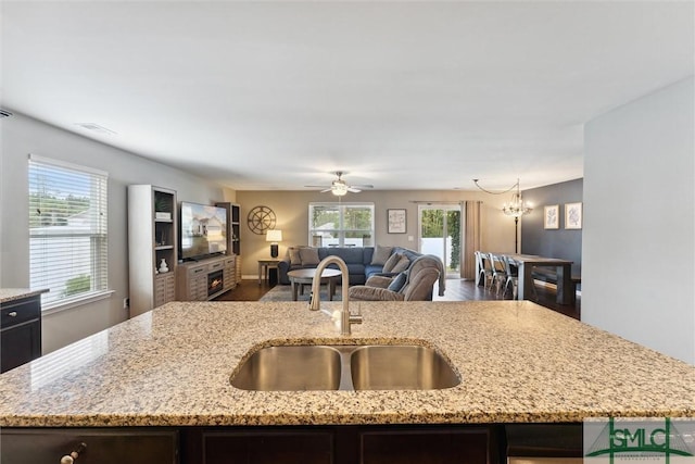 kitchen with light stone countertops, sink, ceiling fan with notable chandelier, and an island with sink