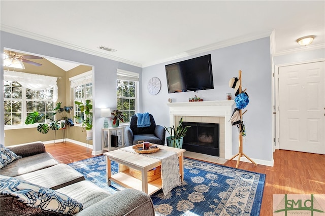 living room with wood-type flooring, a tile fireplace, ceiling fan, and crown molding