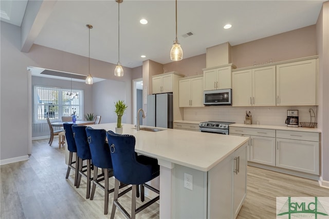 kitchen featuring sink, appliances with stainless steel finishes, light wood-type flooring, a kitchen island with sink, and pendant lighting