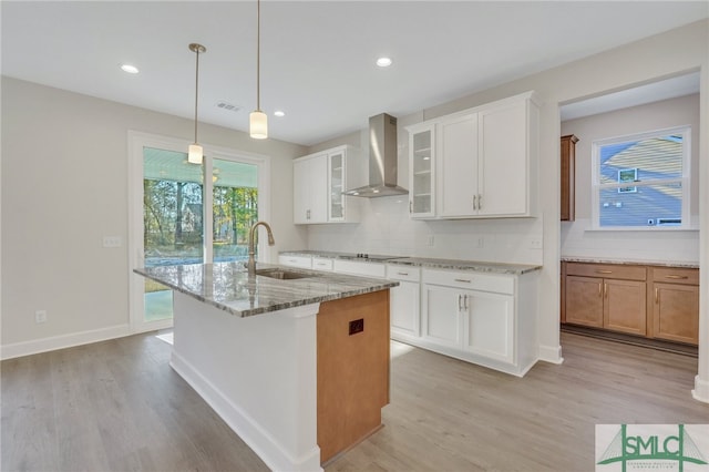 kitchen featuring light stone counters, wall chimney exhaust hood, sink, white cabinets, and an island with sink