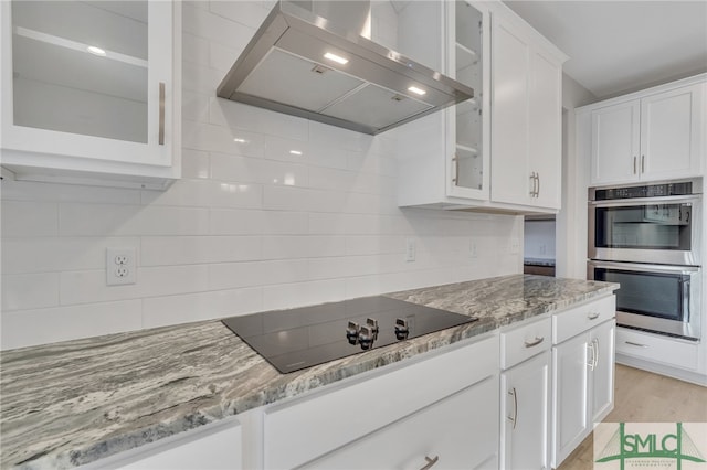 kitchen with white cabinets, wall chimney range hood, black electric cooktop, double oven, and light stone counters