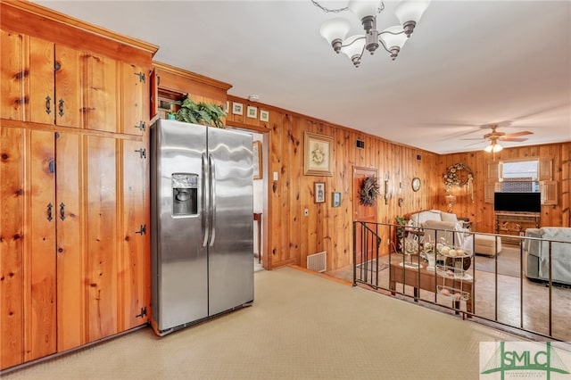 kitchen with light carpet, stainless steel fridge with ice dispenser, ceiling fan with notable chandelier, and wood walls