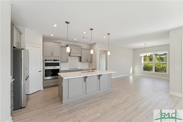 kitchen featuring appliances with stainless steel finishes, premium range hood, sink, light hardwood / wood-style floors, and an island with sink