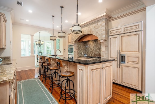 kitchen with ornate columns, dark stone countertops, an island with sink, wood-type flooring, and decorative light fixtures