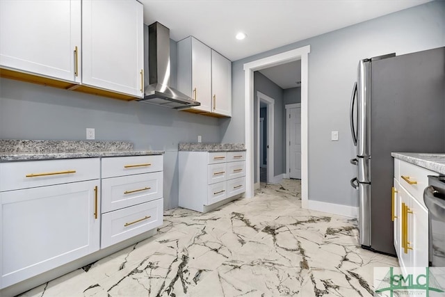 kitchen featuring wall chimney range hood, stainless steel refrigerator, white cabinets, and light stone counters