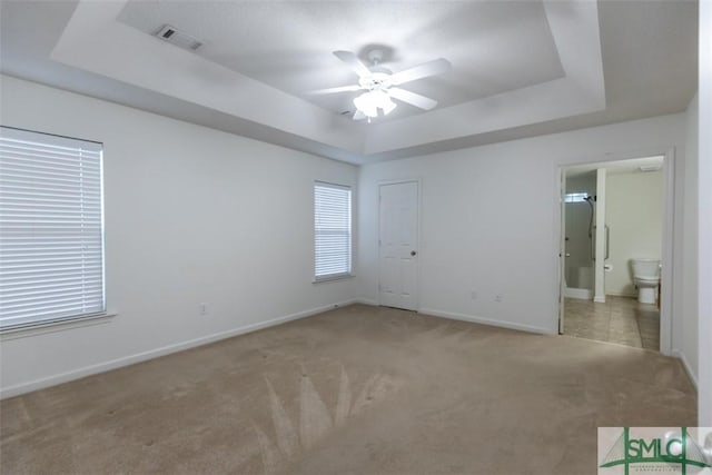 unfurnished bedroom with a tray ceiling, ensuite bath, light carpet, and ceiling fan