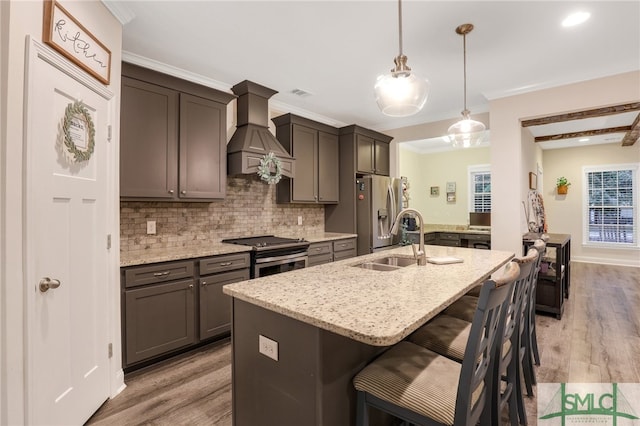 kitchen featuring stainless steel appliances, light hardwood / wood-style flooring, premium range hood, pendant lighting, and a center island with sink