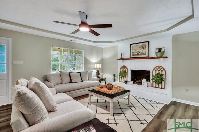 living room featuring a tray ceiling, a brick fireplace, ceiling fan, and light wood-type flooring