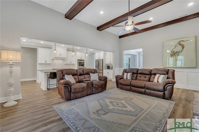 living room with ceiling fan, a towering ceiling, beamed ceiling, and light wood-type flooring