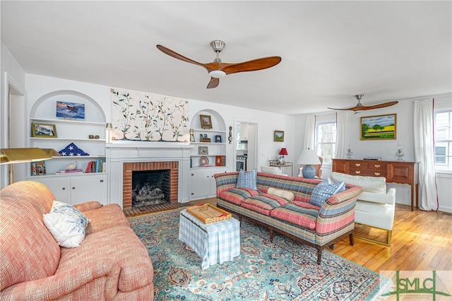 living room featuring built in shelves, ceiling fan, a fireplace, and light hardwood / wood-style flooring