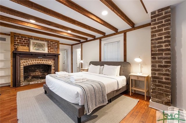 bedroom with beamed ceiling, wood-type flooring, and a brick fireplace