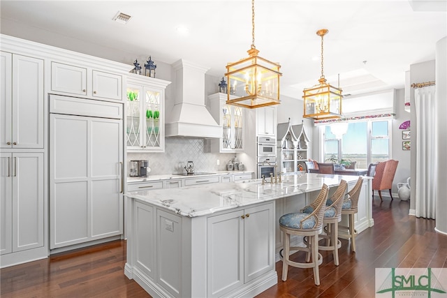 kitchen featuring pendant lighting, premium range hood, a center island with sink, white cabinets, and dark hardwood / wood-style flooring