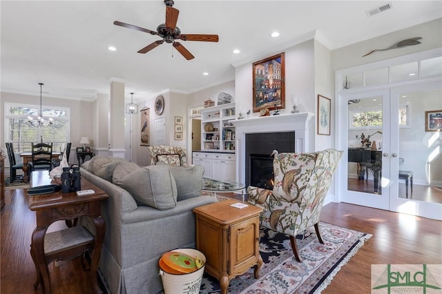 living room featuring french doors, dark wood-type flooring, crown molding, a tiled fireplace, and ceiling fan with notable chandelier