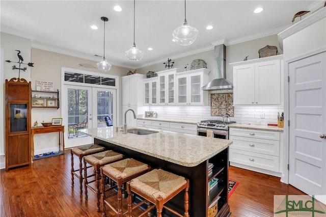 kitchen with french doors, wall chimney exhaust hood, sink, a center island with sink, and white cabinetry