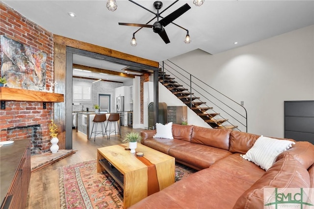 living room with ceiling fan, beam ceiling, light hardwood / wood-style floors, brick wall, and a brick fireplace