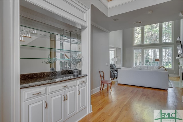 bar featuring dark stone countertops, crown molding, a towering ceiling, light hardwood / wood-style floors, and white cabinets