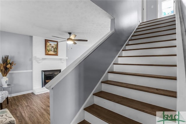 staircase with ceiling fan, wood-type flooring, and a brick fireplace