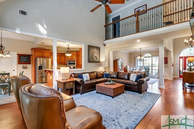 living room featuring a towering ceiling, ceiling fan with notable chandelier, decorative columns, wood-type flooring, and crown molding