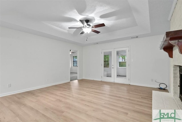 spare room with light hardwood / wood-style flooring, ceiling fan, a tray ceiling, a fireplace, and french doors